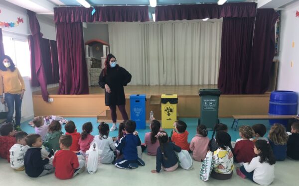 Niños sentados en un taller de reciclaje con instructora y contenedores de colores en un escenario.