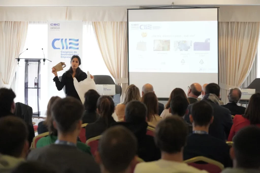 Mujer presenta sobre sostenibilidad frente a audiencia en congreso, sujetando una bolsa y papel.