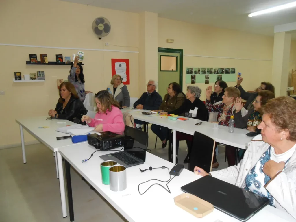 Personas participando en una reunión educativa en una sala con mesas y portátiles. Una persona sostiene un libro.