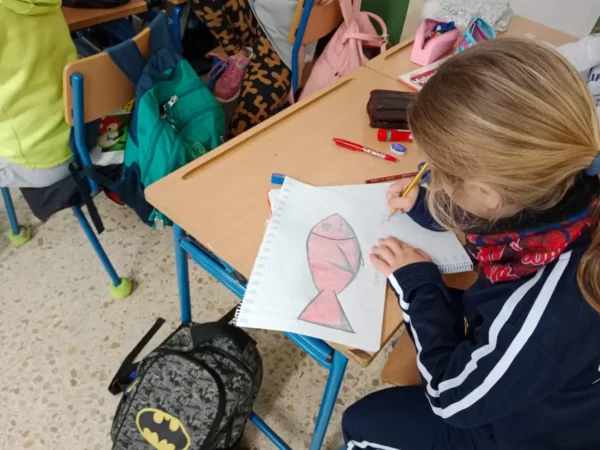 Niña dibuja un atún rojo sobre papel en un aula. Hay mochilas y útiles escolares sobre el escritorio.