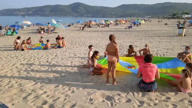 Niños jugando con una tela colorida en la arena de una playa concurrida, con montañas al fondo.