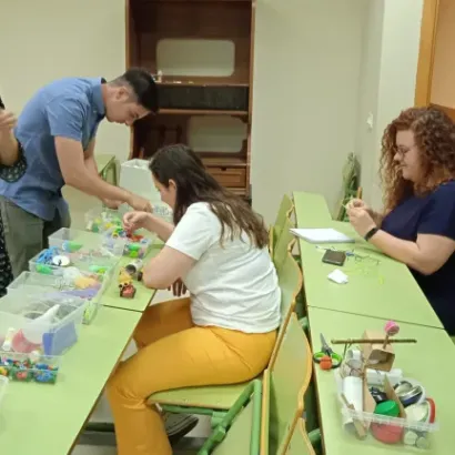 Personas participando en un taller de manualidades en un aula, usando materiales diversos sobre mesas verdes.