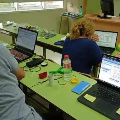 Personas trabajando en una sala de clase con laptops en escritorios verdes, rodeados de material de oficina.