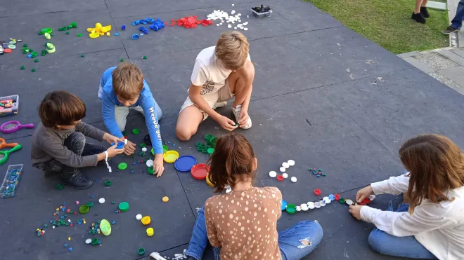 Niños jugando y creando figuras con tapas de plástico de colores sobre el suelo en un ambiente al aire libre.