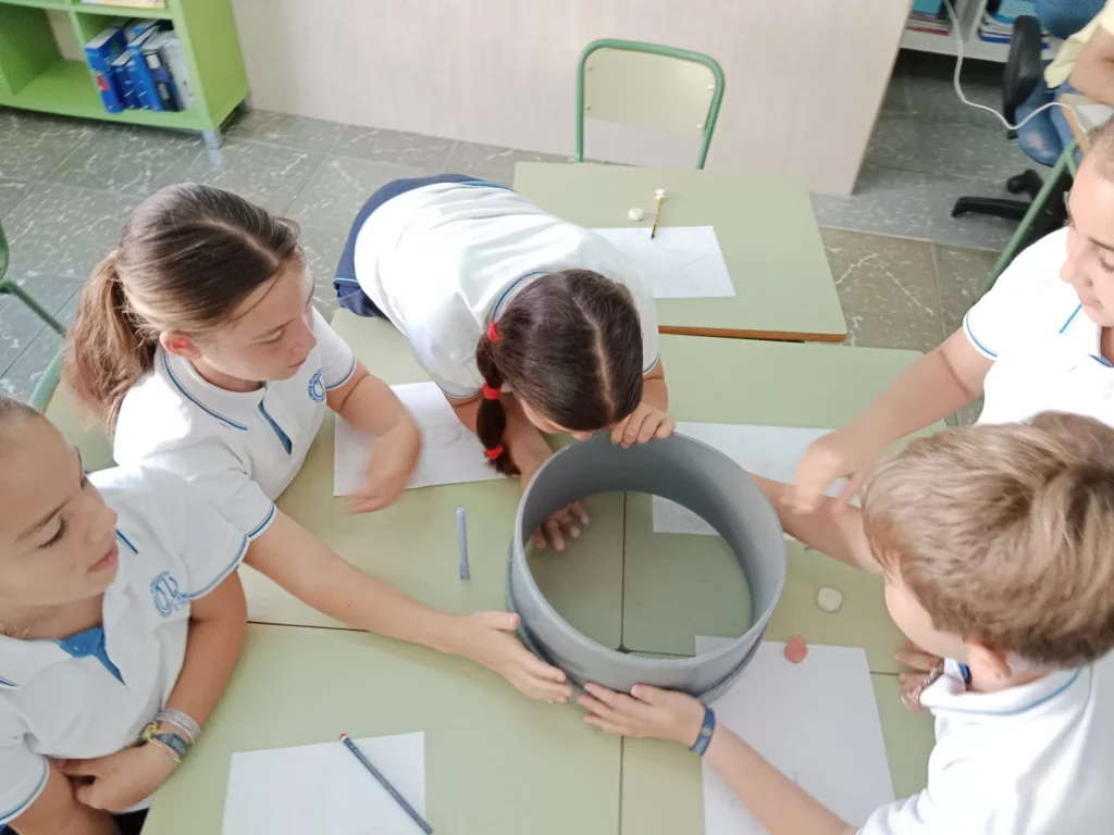 Estudiantes colaboran en clase, observando un cilindro gris sobre una mesa con papeles y lápices.