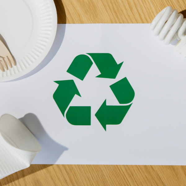Símbolo de reciclaje verde sobre papel, rodeado de platos, utensilios de papel, y una bombilla en una mesa de madera.