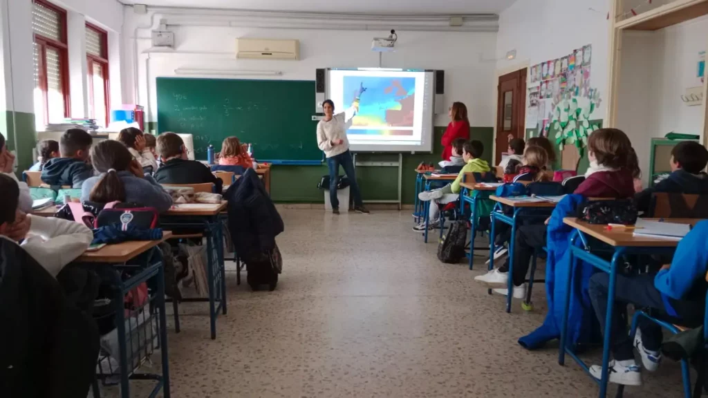 Clase en un aula con estudiantes sentados, un docente explicando frente a una pantalla proyectada en la pizarra.