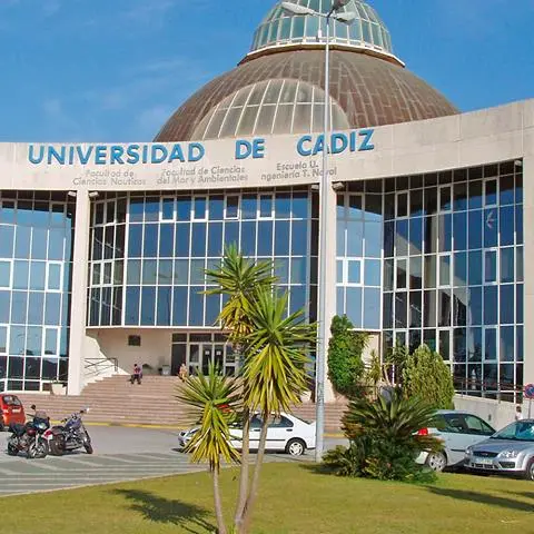 Fachada de la Universidad de Cádiz, con cúpula de vidrio y plantas en el frente, bajo un cielo despejado.
