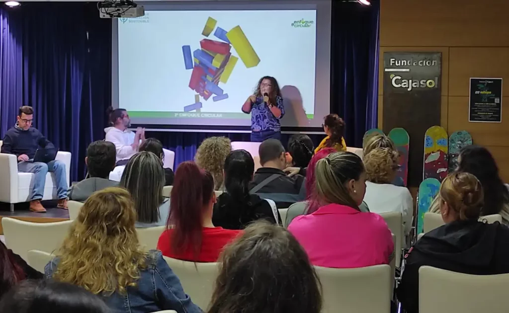 Una presentación en un auditorio donde una persona habla frente a una pantalla con figuras coloridas.