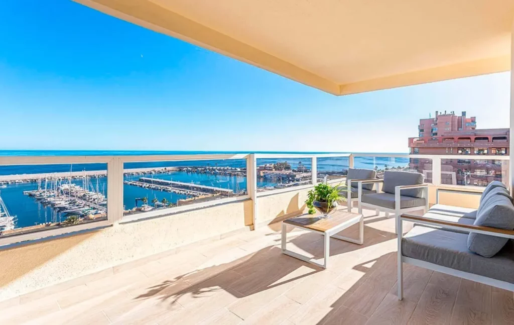 Balcón con vista al puerto deportivo, muebles de exterior y plantas bajo un cielo azul despejado.