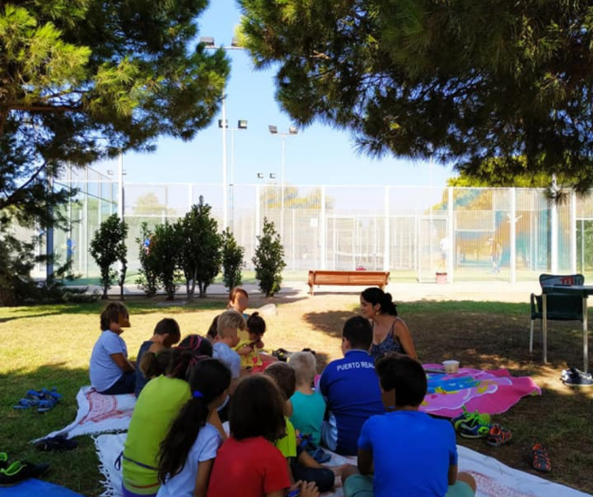 Grupo de niños y una adulta sentados en un césped a la sombra, asistiendo a un taller al aire libre.