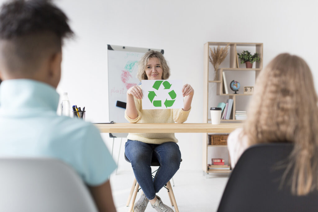 Persona enseñando reciclaje con un cartel de símbolo de reciclaje a dos estudiantes en un aula.