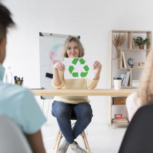 Persona enseñando reciclaje con un cartel de símbolo de reciclaje a dos estudiantes en un aula.