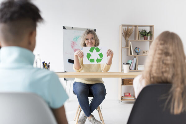 Persona enseñando reciclaje con un cartel de símbolo de reciclaje a dos estudiantes en un aula.