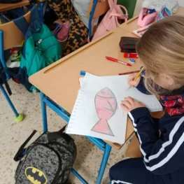 Niña dibuja un atún rojo sobre papel en un aula. Hay mochilas y útiles escolares sobre el escritorio.
