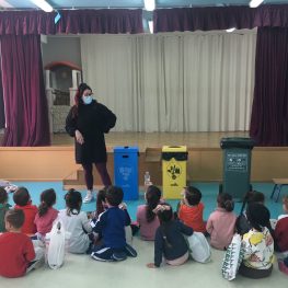 Niños sentados en un taller de reciclaje con instructora y contenedores de colores en un escenario.